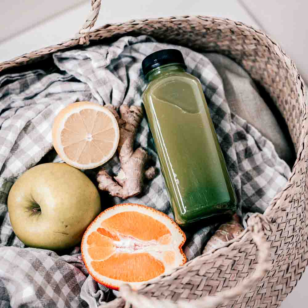 A woven basket containing fresh green juice, apples, and citrus fruits for the Guided Liver and Gallbladder Cleanse by Emily Schwarze.